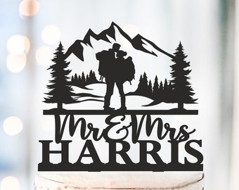 May include: A black cake topper featuring a silhouette of a couple with backpacks in front of mountains and trees. The words "Mr & Mrs HARRIS" are below the silhouette. The cake is a multi-layered cake with a red base.