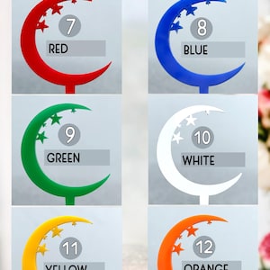 May include: Acrylic cake toppers in the shape of a crescent moon with stars. The toppers are available in red, blue, green, white, yellow, and orange. The numbers 7 through 12 are printed on the toppers.