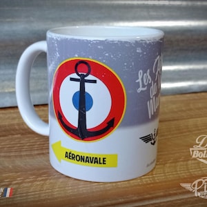 Könnte beinhalten: Weißer Keramikbecher mit einem Design mit einem schwarzen Anker in einem rot-weißen Kreis mit blauem Zentrum. Der Becher trägt die Aufschrift "AÉRONAVALE" und "Les As de la Marine". Der Becher trägt auch die Aufschrift "FABRICATION ARTISANALE".