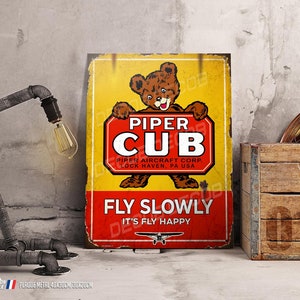 Peut inclure: Une plaque en métal de style vintage représentant un ours en dessin animé portant une casquette de pilote et tenant une pancarte sur laquelle est inscrit "Piper Cub". La plaque est jaune avec des accents rouges et blancs et porte le texte "Fly Slowly, It's Fly Happy" en bas.
