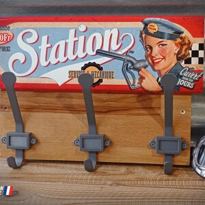Può includere: Appendiabiti da parete in legno con tre ganci in metallo. Il portabiti presenta un'insegna in stile vintage con una donna che tiene in mano una pompa di benzina, la parola "Station" e il testo "Service & Mecanique".