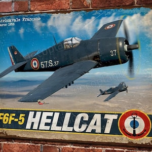 Könnte beinhalten: Ein Vintage-Metallschild mit einem blauen und weißen F6F-5 Hellcat-Kampfflugzeug mit dem Text "Aéronavale Française Maroc 1956" und "57.S.37" auf dem Flugzeug. Das Schild enthält auch den Text "F6F-5 HELLCAT" und ein rundes Emblem mit einem weißen Anker auf blauem Hintergrund.