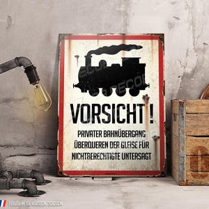 Op de afbeelding: Een rechthoekig metalen bord met een rode rand en het woord "VORSICHT!" in grote witte letters. Een zwart silhouet van een trein staat boven de tekst. Het bord bevat ook Duitse tekst en staat naast een houten kist en een pijplamp.