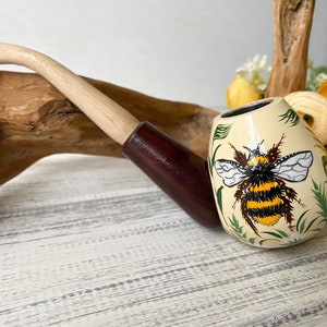 May include: A hand-painted wooden pipe with a brown stem and a white bowl. The bowl is decorated with a yellow and black bumblebee surrounded by green leaves.