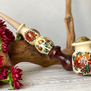 May include: A hand-painted wooden pipe with a floral design in shades of orange, green, and brown. The pipe has a cream-colored base and a brown wooden stem.