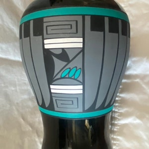 May include: Black ceramic vase with a geometric design in shades of gray, teal, and white. The vase has a wide base and a narrow neck.