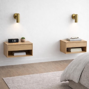 May include: Two light wood floating bedside tables with a single drawer and an open shelf, each mounted on a white wall beneath a cylindrical brass wall light. The left bedside table holds a black digital clock and a small succulent plant.