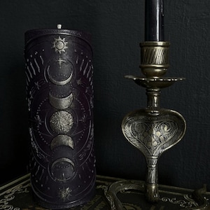 May include: A black candle with a silver moon and sun design on a dark background. The candle is sitting on a book with the words "Magic Spells" printed on the cover. A gold metal candle holder shaped like a snake is in the background.
