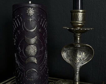May include: A black candle with a silver moon and sun design on a dark background. The candle is sitting on a book with the words "Magic Spells" printed on the cover. A gold metal candle holder shaped like a snake is in the background.