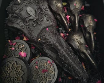 May include: Black skull-shaped candles with intricate details, including a fleur-de-lis design, surrounded by black and pink rose petals.