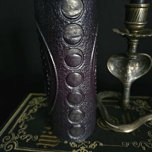 May include: A black candle with a textured surface and silver moon phases embossed on the side. The candle is sitting on a black book with gold accents.