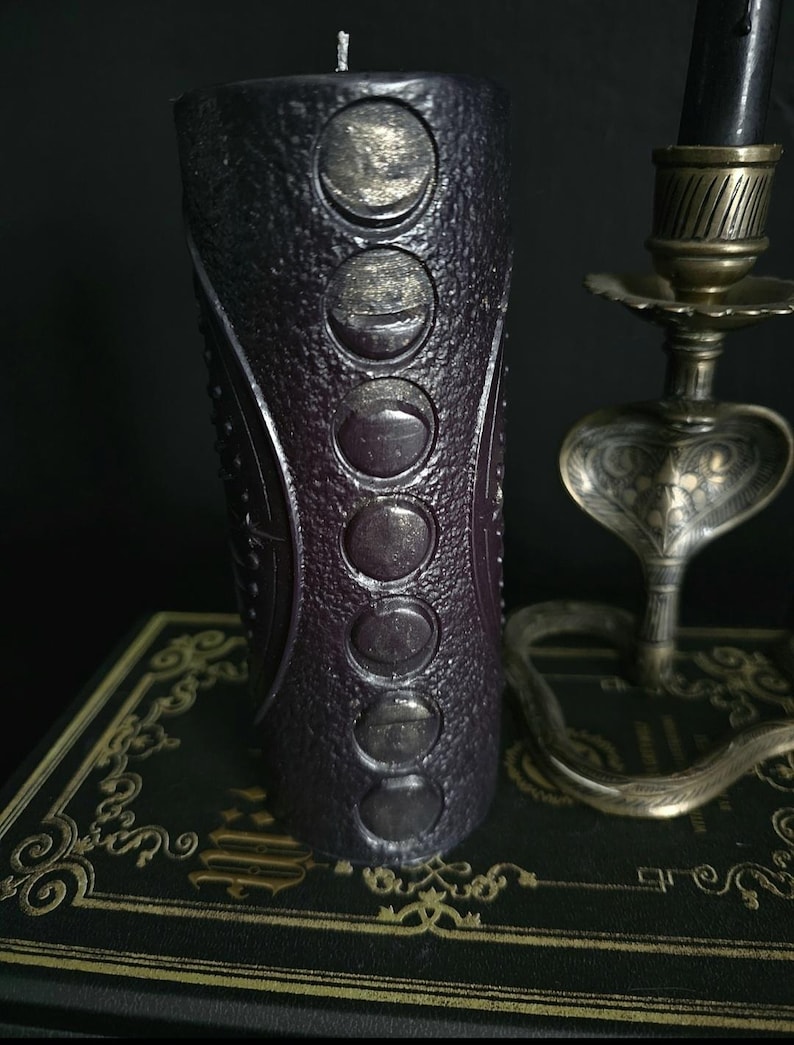 May include: A black candle with a textured surface and silver moon phases embossed on the side. The candle is sitting on a black book with gold accents.