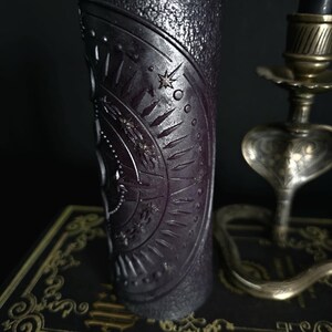 May include: A black pillar candle with a textured surface and a circular design featuring a star and other symbols. The candle is sitting on a book with a gold design.