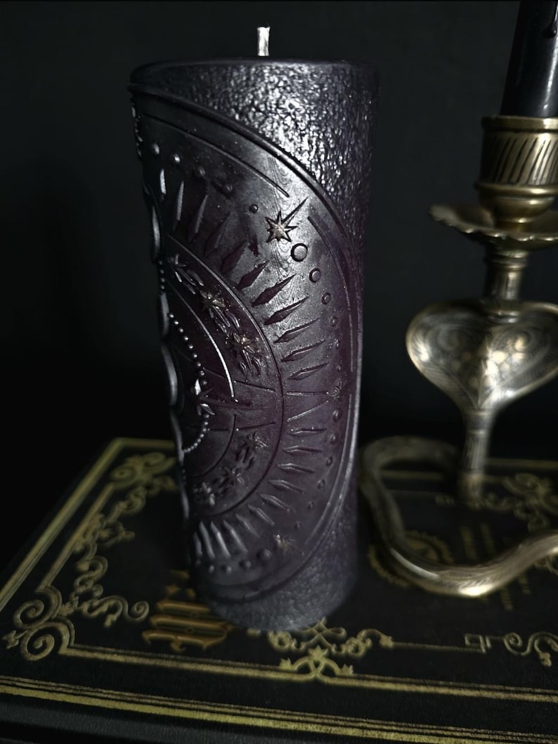 May include: A black pillar candle with a textured surface and a circular design featuring a star and other symbols. The candle is sitting on a book with a gold design.