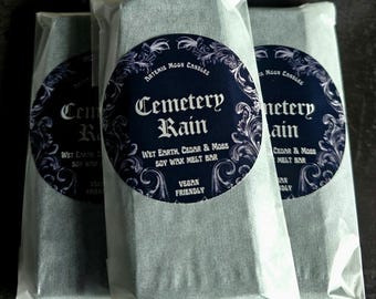 May include: Three soy wax melt bars in white paper packaging. Each bar has a dark blue circular label with the text "Cemetery Rain" and "Wet Earth, Cedar & Moss Soy Wax Melt Bar". The labels also include the words "Artemis Moon Candles" and "Vegan Friendly".
