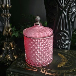 May include: A pink glass jar with a diamond pattern and a silver lid. The jar is round and has a matching lid with a small, decorative knob. The jar is sitting on a dark surface.