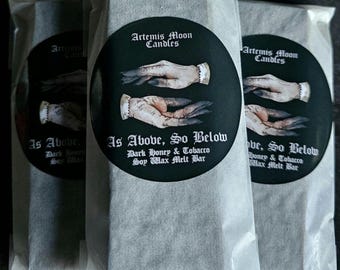 May include: Three white paper bags with black text and a black and white illustration of hands. The text on the bags reads "Artemis Moon Candles" and "As Above, So Below Dark Honey & Tobacco Soy Wax Melt Bar".