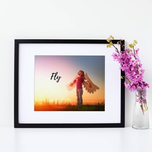Pode incluir: Uma impressão emoldurada com uma foto de uma criança usando asas de papel e olhando para o pôr do sol. A palavra "Fly" está escrita em texto preto na impressão.