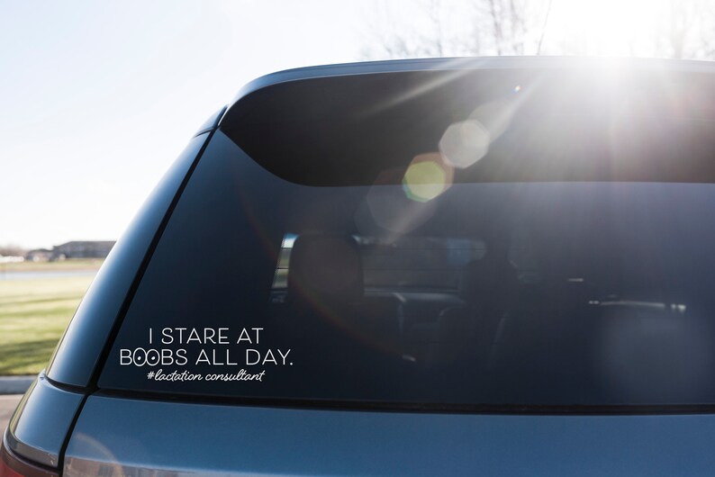 May include: A white decal on a car window reads "I STARE AT BOOBS ALL DAY. #lactation consultant". The text is in a sans-serif font. The car is a dark blue color, and the background is a bright, sunny day.
