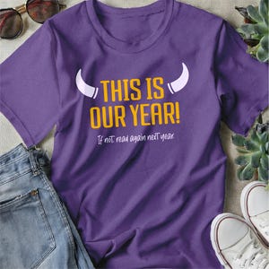 May include: A purple t-shirt with the text "THIS IS OUR YEAR!" in yellow and white. Below the main text is the phrase "If not, read again next year." The shirt is paired with blue jeans and white sneakers.