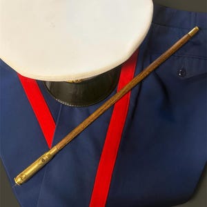 May include: A white military-style peaked cap with a black brim, placed atop blue trousers featuring a prominent red vertical stripe. A long, thin wooden swagger stick with gold-colored metal ends, one featuring an engraved chevron insignia.