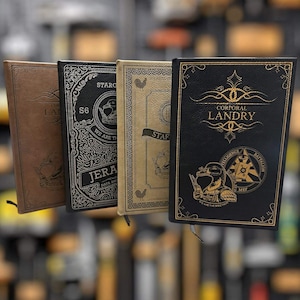 May include: Four leather-bound journals with embossed designs. The journals are in shades of brown, black, and tan. One journal reads "CORPORAL LANDRY" with a gold emblem. Other journals have text and illustrations, including "STARG" and "JERA DATA ARC".
