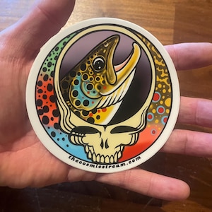 May include: A round sticker featuring a brown trout with an open mouth, set against a purple background. The fish is surrounded by a colorful, rainbow-like border and a stylized skull design. The text "thecosmicstream.com" is at the bottom.