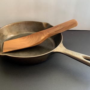 May include: A wooden spatula resting in a cast iron skillet. The spatula is made of light brown wood and has a curved shape. The skillet is dark grey and has a handle.