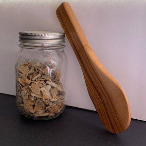 May include: A glass jar filled with brown, irregularly shaped pieces of food, with a silver lid. A wooden spoon with a curved handle is next to the jar.