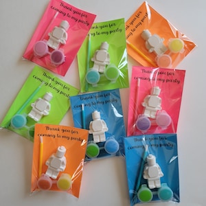 May include: Eight favor bags with a white Lego figurine, paint pots, and a paintbrush. The bags are decorated with neon pink, green, orange, and blue backgrounds. The text on the bags reads "Thank you for coming to my party".