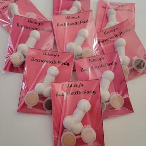 May include: A set of seven pink and white party favors with the text "Hilary's Bachelorette Party" printed on each. Each favor includes a white plastic figurine, two small containers of paint, and a small paintbrush.