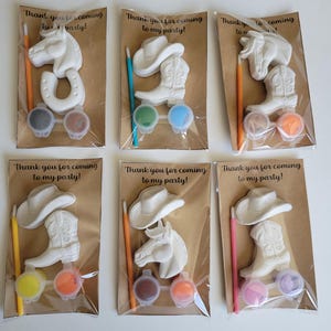 May include: Six party favor sets with a cowboy theme. Each set includes a white plaster horse head, horseshoe, cowboy hat, or boot, paint pots, and a paintbrush. The sets are packaged in clear plastic bags with a brown paper backing. Text on the packaging reads "Thank you for coming to my party!"