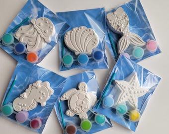 May include: Six individual paint-your-own ceramic ornaments with paint pots and brushes. The ornaments feature sea-themed designs, including a seashell, a starfish, a turtle, a seahorse, a jellyfish, and a mermaid tail.