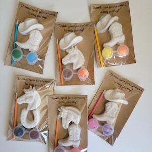 May include: Six party favor sets with cowboy-themed figurines, paint pots, and paintbrushes. Each set includes a cowboy hat, a boot, a horse head, or a horseshoe. The text "Thank you for coming to my party!" is printed on the packaging.