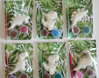May include: Six individual clear plastic bags, each containing a white plaster dinosaur figurine, a small paintbrush, and two small circular paint pots. The backing card inside each bag features a green tropical leaf pattern design.