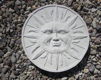 Puede incluir: Una escultura de sol de hormigón blanco con una cara sonriente y rayos que emanan de la cabeza. La escultura está sentada sobre un lecho de pequeñas piedras grises y marrones.