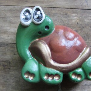 Puede incluir: Una figurita de tortuga de cerámica verde y marrón con grandes ojos blancos. La tortuga tiene un borde dorado alrededor de su caparazón.