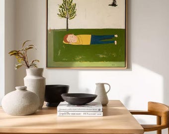 May include: A square framed painting depicts a person with reddish hair wearing a yellow shirt and blue pants lying on a green lawn under a tree. A small black figure is in the upper right corner. The painting is displayed above a wooden table.