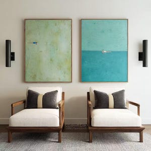 Op de afbeelding: Twee rechthoekige ingelijste schilderijen hangen boven twee houten fauteuils met crèmekleurige kussens en donkergrijze sierkussens. Het linker schilderij heeft een lichtgroene achtergrond met een klein figuurtje, het rechter toont een blauwe zee.