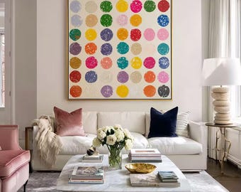 May include: A large square wall art piece featuring a grid of colorful, textured circles in shades of red, blue, yellow, green, pink, orange, and purple against a cream background, enclosed in a thin gold frame and displayed above a white sofa.
