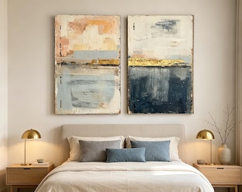 May include: Two rectangular abstract paintings featuring textured layers of cream, soft peach, light blue, deep navy, and metallic gold leaf accents. The artworks are displayed side-by-side above a bed with neutral bedding and two bedside lamps.