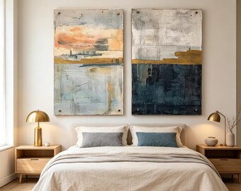 May include: Two large rectangular abstract paintings mounted on a wall above a bed. The left painting features soft blue, peach, and gold tones. The right painting displays a bold contrast of dark navy blue, white, and a horizontal gold band.