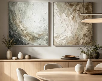 May include: Two square canvas paintings featuring heavy impasto texture in shades of white, beige, and charcoal gray. The artwork displays swirling, abstract brushstrokes and thick paint application, mounted side-by-side on a neutral wall.