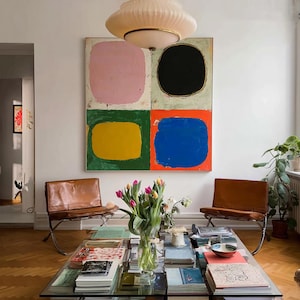 Può includere: Un interno con un grande dipinto astratto con quattro blocchi colorati: rosa, nero, giallo e blu. Due sedie in pelle marrone affiancano un tavolino con piano in vetro, libri e un vaso di tulipani. Un grande lampadario color crema pende dal soffitto.