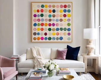 May include: A large square wall art piece featuring a grid of colorful painted circles in shades of red, blue, yellow, green, pink, and brown on a cream background, framed in a thin gold border, displayed above a white sofa in a living room.