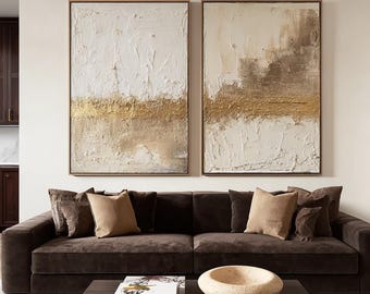 May include: Two rectangular wall art canvases featuring heavy white and beige impasto texture with a horizontal band of metallic gold paint across the center. The paintings are framed in thin wood and displayed side by side on a plain wall.