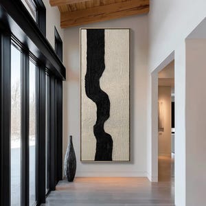 Puede incluir: Una pintura abstracta vertical grande con una forma negra similar a un río sobre un fondo texturizado blanquecino. La obra de arte está enmarcada en un borde delgado de color dorado. Un jarrón alto y oscuro está en el suelo frente a una ventana grande.