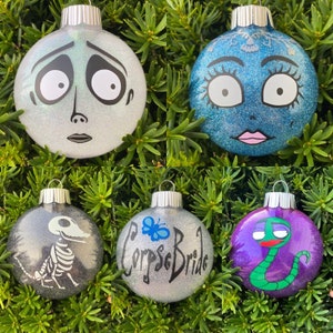 May include: Five round Christmas ornaments with glitter and hand-painted designs. The ornaments feature characters from the movie Corpse Bride. One ornament is silver with a black skeleton, one is silver with a blue butterfly and the text "Corpse Bride", one is purple with a green snake, one is silver with a white face and black eyes, and one is blue with a white face and black eyes.