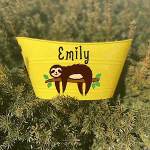 May include: A bright yellow metal bucket with a black cursive name "Emily" and a brown sloth illustration resting on a branch with green leaves. The bucket has a rounded shape and is sitting in front of green foliage.