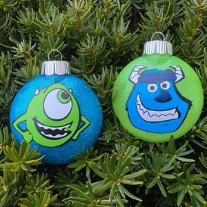 May include: Two Christmas ornaments featuring characters from the movie Monsters Inc. One ornament is green with a blue glitter background and features Sulley, the blue monster with one eye. The other ornament is blue with a green glitter background and features Mike, the green one-eyed monster.
